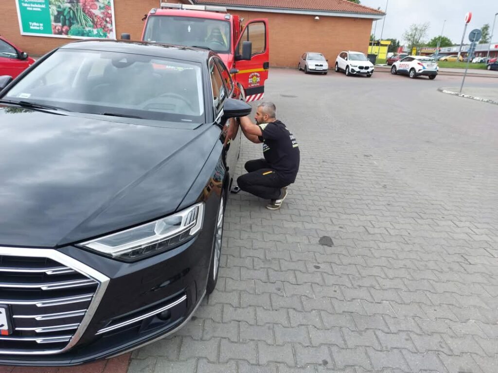 Mężczyzna wykonujący awaryjne otwieranie samochodu na parkingu przy czarnym Audi.