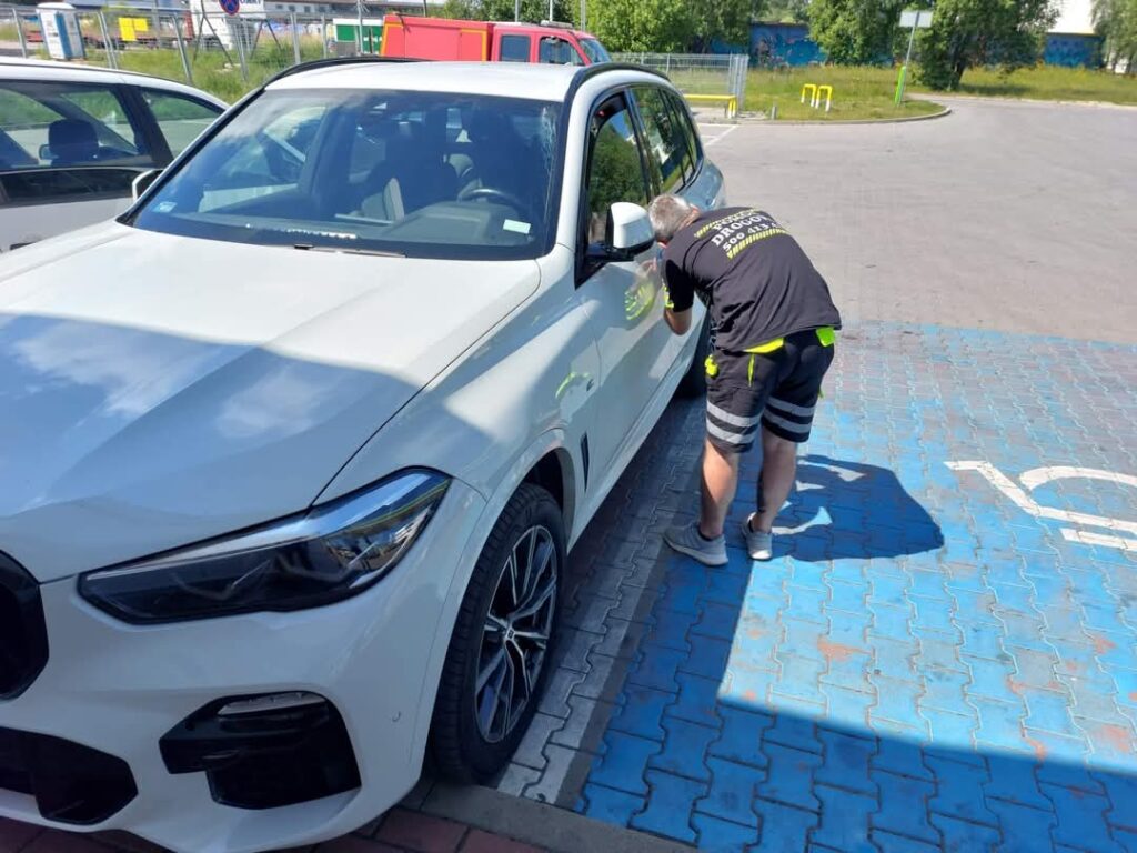 Awaryjne otwieranie drzwi białego BMW na miejscu parkingowym dla osób niepełnosprawnych.