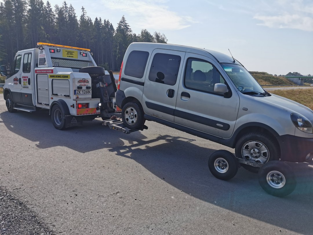 Srebrny van holowany przez lawetę na poboczu trasy ekspresowej w słoneczny dzień.