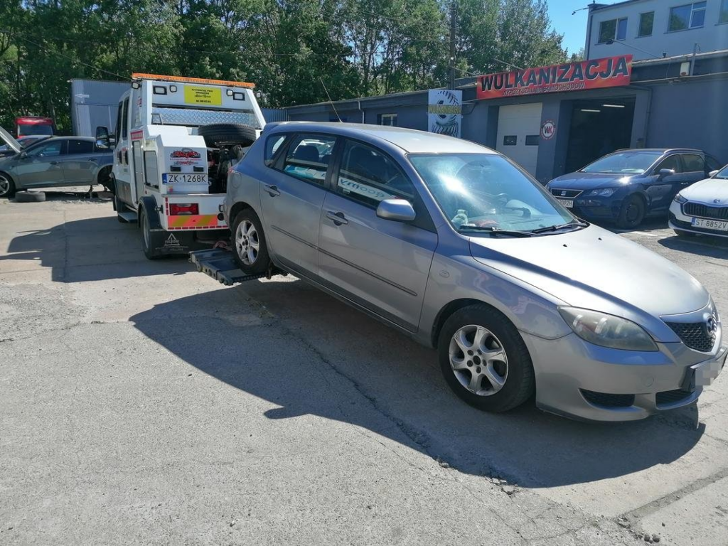 Laweta transportująca szare kombi na tle miejskiego parkingu i ulicznego ruchu.