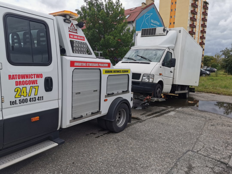 Pomoc drogowa holująca biały samochód dostawczy z uszkodzonym przednim kołem.