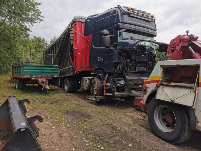 Uszkodzony czarny ciągnik siodłowy z naczepą w trakcie akcji ratowniczej poza miastem.
