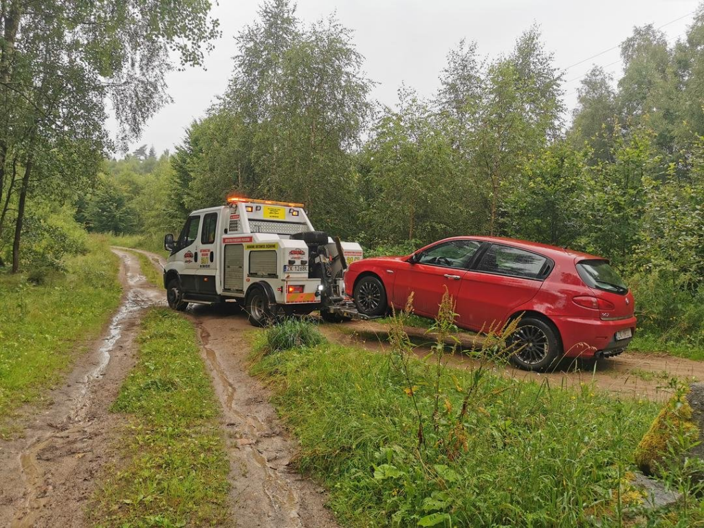 Czerwony samochód osobowy holowany z leśnej, błotnistej drogi przez lawetę.