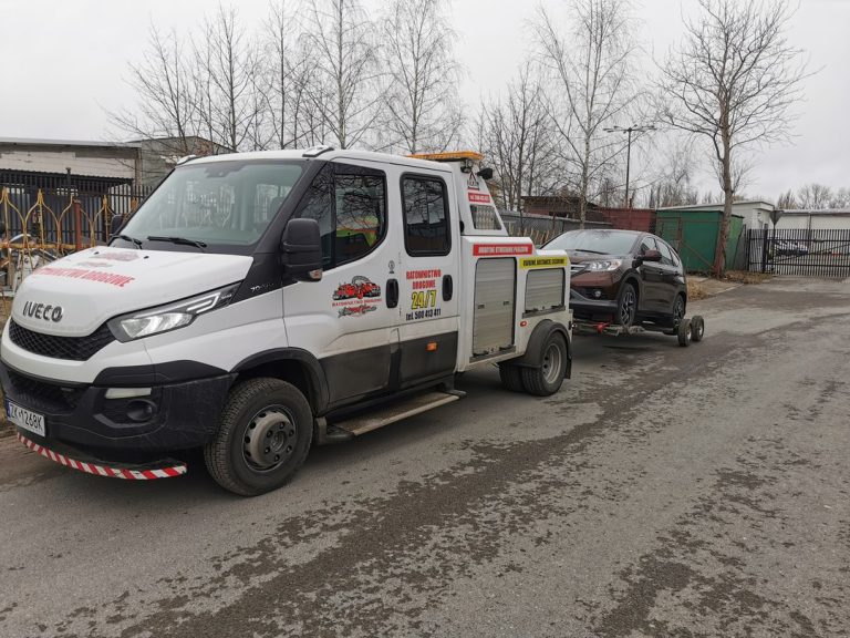 Pojazd pomocy drogowej transportujący brązowy samochód na obrzeżach miasta.
