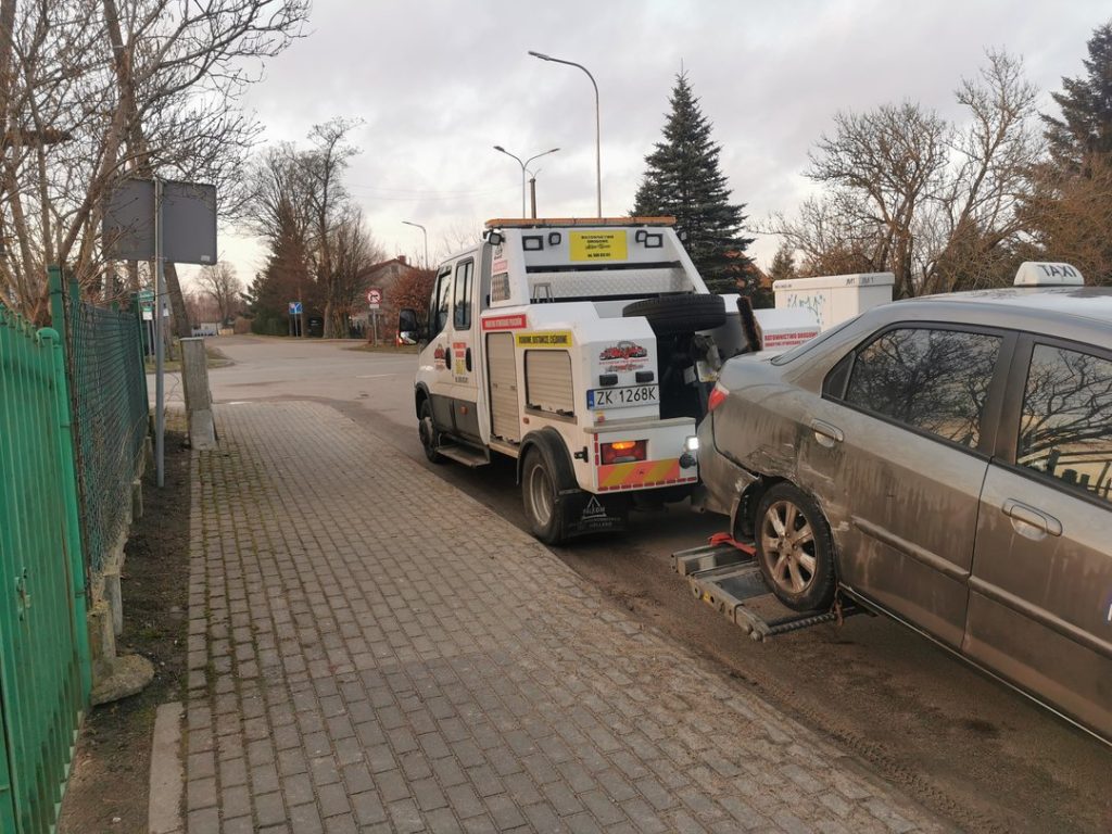 Holowanie szarego auta osobowego z widocznymi uszkodzeniami tylnej osi na osiedlowej ulicy.